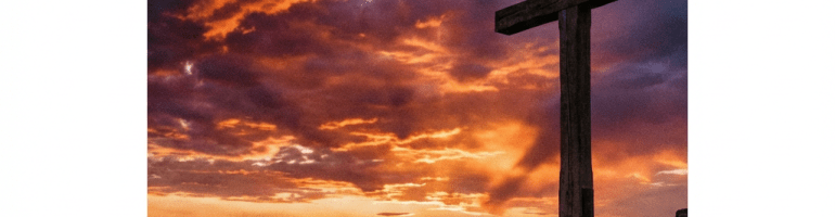A wooden cross on a rocky hill under a dramatic sunset sky with valley fog.